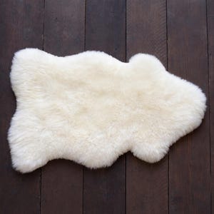 May include: A cream-colored sheepskin rug with a unique, irregular shape. The rug is made of soft, fluffy wool and is placed on a dark brown wooden surface. The rug's texture and color create a cozy and inviting feel.