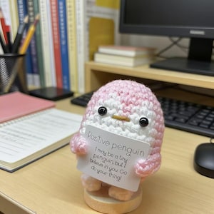 May include: A handmade pink and white crocheted penguin figurine holding a small card. The card reads "Positive penguin" with an inspirational message. The penguin is on a wooden base.