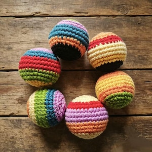 May include: Six colorful, crocheted balls arranged in a circle. Each ball features a unique combination of vibrant colors, including red, blue, green, orange, and yellow. The balls are set against a rustic wooden background.