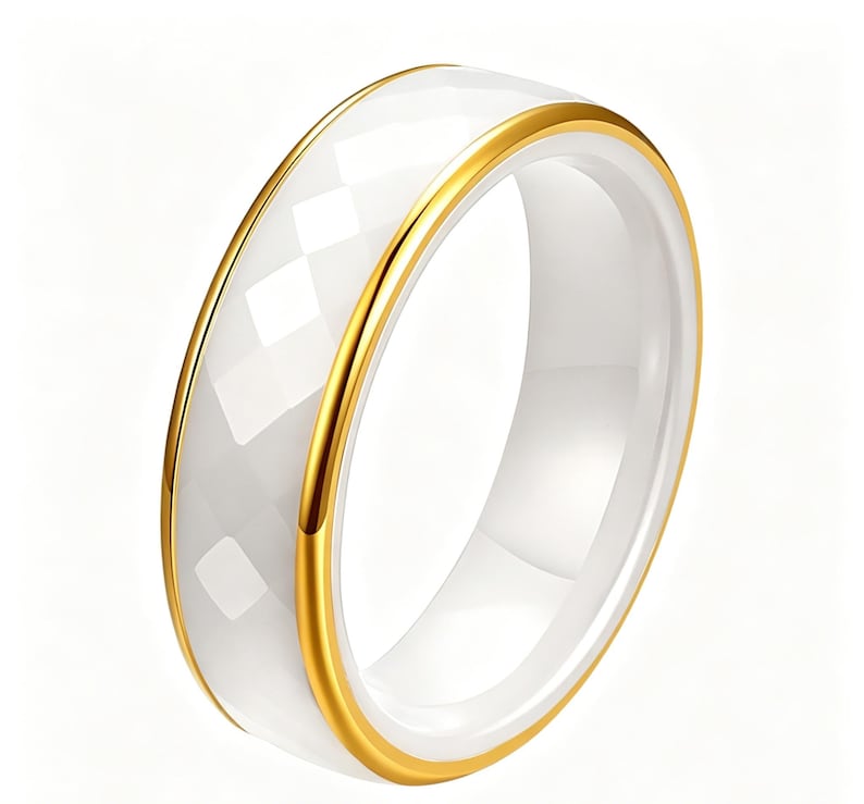 Op de afbeelding: Een witte keramische ring met een facetgeslepen buitenband en een gepolijste binnenband. De ring heeft een goudkleurige metalen rand aan beide randen. Een eenvoudig, elegant sieraad.