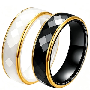 Op de afbeelding: Twee ringen, &eacute;&eacute;n wit en &eacute;&eacute;n zwart, met een facetontwerp. Beide ringen hebben een goudkleurige band langs de randen. De witte ring staat links en de zwarte ring rechts. De ringen staan tegen een witte achtergrond.