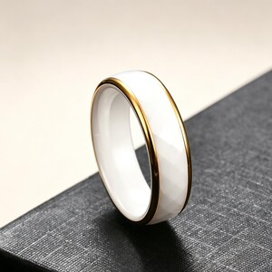 Op de afbeelding: Een witte keramische ring met een goudkleurige band. De ring heeft een facetgeslepen oppervlak en een gepolijste afwerking. De gouden band voegt een vleugje elegantie toe aan het eenvoudige ontwerp. Deze ring is een sieraad.