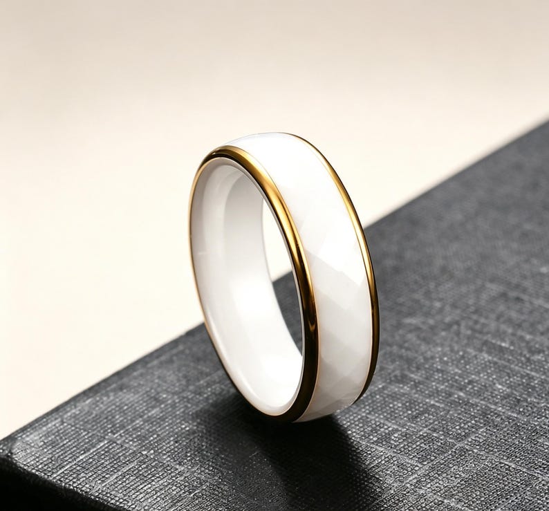 Op de afbeelding: Een witte keramische ring met een goudkleurige band. De ring heeft een facetgeslepen oppervlak en een gepolijste afwerking. De gouden band voegt een vleugje elegantie toe aan het eenvoudige ontwerp. Deze ring is een sieraad.