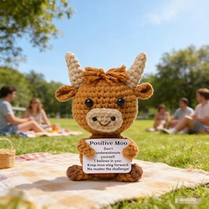 May include: A crocheted brown cow with tan horns and a smiling face. The cow holds a sign that reads "Positive Moo" with an inspirational message. The cow is sitting on a blanket in a park with people in the background.