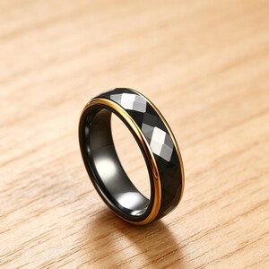 Op de afbeelding: Een zwarte ring met een facetgeslepen, geometrisch ontwerp en een goudkleurige band. De ring wordt getoond op een houten oppervlak, wat de gepolijste afwerking en moderne esthetiek benadrukt. Het ontwerp van de ring heeft een reeks schuine facetten die het licht vangen.