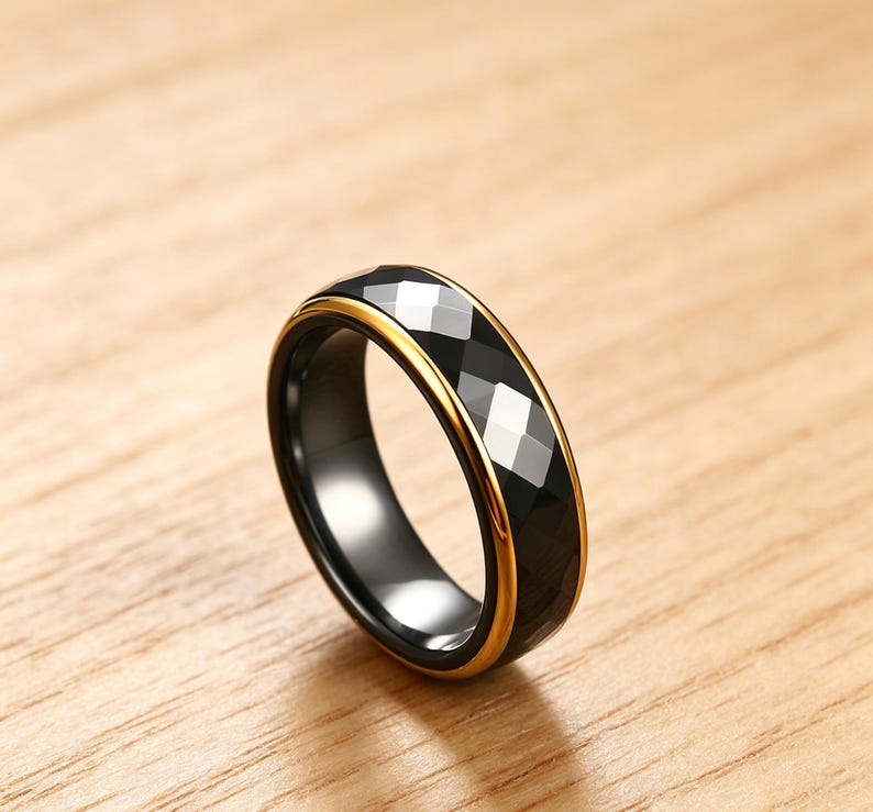 Op de afbeelding: Een zwarte ring met een facetgeslepen, geometrisch ontwerp en een goudkleurige band. De ring wordt getoond op een houten oppervlak, wat de gepolijste afwerking en moderne esthetiek benadrukt. Het ontwerp van de ring heeft een reeks schuine facetten die het licht vangen.
