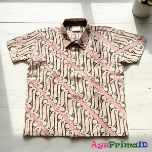 May include: A short-sleeved shirt with a pink and black batik pattern. The shirt has a pocket on the left chest and a collar. The text "AyuPrimaID" is visible at the bottom right.