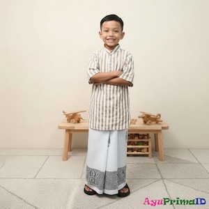 May include: A young person wearing a striped shirt and white sarong with a black and white patterned border. The shirt has vertical brown and white stripes. The sarong is white with a black and white patterned border. The person is standing in front of a wooden table with toy airplanes.