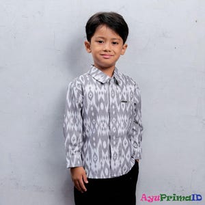 May include: A young person wearing a long-sleeved button-down shirt with a gray and white ikat pattern. The shirt has a collared neckline and a small pocket on the chest. The person is also wearing black pants.