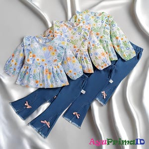 Peut inclure: Ensemble de vêtements pour enfants composé de hauts à imprimé floral avec des bords à volants et des manches longues, associés à des pantalons en jean bleu. Les pantalons ont des bas évasés et des nœuds roses. Les hauts ont un fond bleu clair avec des fleurs jaunes, oranges et bleues.