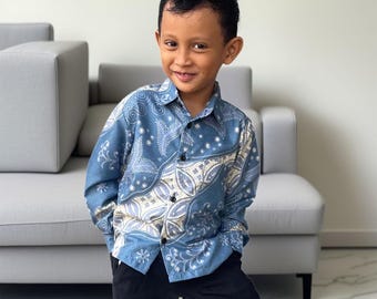 May include: A young person wearing a blue patterned button-down shirt and black pants. The shirt features a detailed batik design in shades of blue, white, and beige. The image is taken indoors with a neutral background.