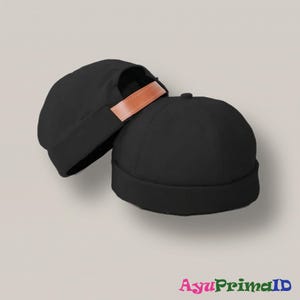 Puede incluir: Dos gorros negros con un detalle de correa de cuero marrón. Los gorros están hechos de una tela suave y tienen una corona redondeada y un borde doblado. Los gorros se muestran sobre un fondo neutro. El texto "AyuPrimaID" es visible en la esquina inferior derecha.