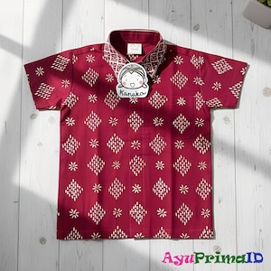 May include: A short-sleeved burgundy shirt with a white geometric pattern of diamonds and flowers. The collar and cuffs have a patterned design. A tag with the name "Kanaka" is attached to the shirt. The shirt is displayed on a light wood surface.