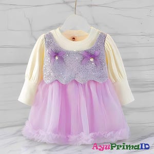 May include: A baby girl's dress with a cream-coloured long-sleeved top and a lavender sequined bodice. The dress features a light purple tulle skirt and is adorned with two small bows. The text "AyuPrimaID" is visible at the bottom.