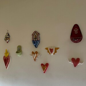 Pode incluir: Uma coleção de decorações de parede em cerâmica em forma de coração em várias cores, incluindo vermelho, dourado e branco. Alguns corações têm asas, enquanto outros apresentam símbolos religiosos. As decorações são montadas numa parede branca.