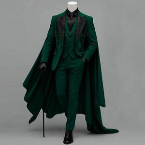 May include: A dark green suit with a matching cape. The suit includes a jacket, waistcoat, and trousers. The jacket has black embroidered detailing on the lapels. The outfit is completed with a black shirt, tie, gloves, and shoes. A black cane is held in the left hand.