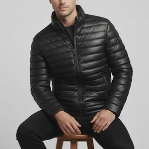 Puede incluir: Una chaqueta acolchada negra con cuello alto y cremallera completa. La chaqueta se lleva con pantalones negros. La persona está sentada en un taburete de madera. La chaqueta parece ser de cuero.