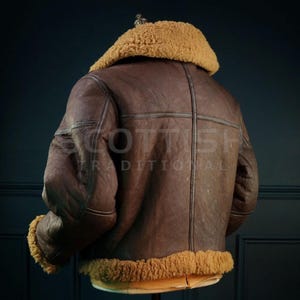Pu&ograve; includere: Giacca in pelle marrone con colletto e finiture in montone beige. La giacca ha un design classico con la schiena cucita a croce e il testo "SCOTTISH TRADITIONAL" nella parte inferiore.
