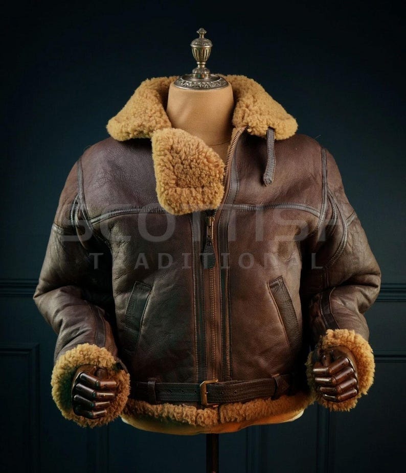 Pu&ograve; includere: Giacca bomber in pelle marrone con colletto e polsini in montone. La giacca ha una chiusura a zip frontale, una cintura in vita e il testo "SCOTTS TRADITIONAL" &egrave; visibile. La giacca &egrave; esposta su un manichino.