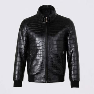 Puede incluir: Chaqueta de cuero negro con textura de piel de cocodrilo. La chaqueta tiene cremallera frontal, cuello y puños y dobladillo de canalé negros. Es una chaqueta estilo bomber.