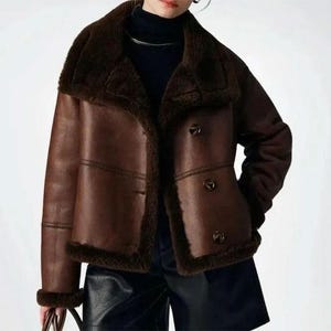 Peut inclure: Veste en peau de mouton marron avec col et garniture en fausse fourrure marron foncé. La veste a une fermeture à boutons et un design classique. La veste est en matière simili cuir.