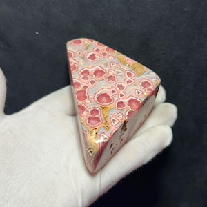 May include: A triangular polished stone specimen featuring a complex pattern of pink, circular inclusions surrounded by grey and white matrix with veins of metallic gold-colored mineral deposits throughout the surface of the rock.