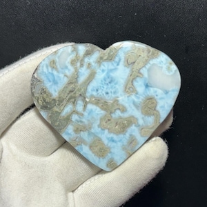May include: A heart-shaped stone with a light blue and tan pattern. The stone is held in a white gloved hand against a black background. The stone has a marbled appearance with a mix of colors and textures.