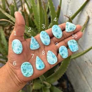 May include: Ten polished Larimar gemstones in various shapes including oval, teardrop, and triangular forms. Each stone displays a distinct light blue color with white marbled patterns. The stones are numbered one through ten in white text.