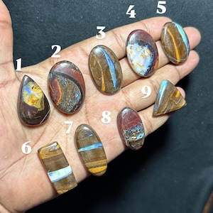 Puede incluir: Una colección de nueve ópalos boulder pulidos etiquetados con los números del uno al nueve. Estas piedras preciosas presentan una matriz de piedra de hierro marrón con destellos iridiscentes de azul, amarillo y blanco. Cada piedra tiene una forma única, incluyendo óvalos, peras y triángulos, mostrando variaciones de color y patrones naturales.