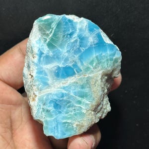 May include: A rough-cut piece of larimar gemstone, showcasing a blend of light blue and white hues with intricate veining. The stone's surface has a textured appearance, with areas of deeper blue and hints of green.