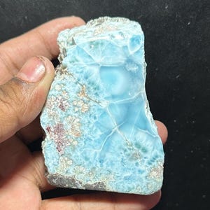 Puede incluir: Un trozo de piedra de larimar en bruto, sostenido en una mano. La piedra presenta un color azul claro con inclusiones blancas y marrones. La superficie tiene un aspecto texturizado con estructuras cristalinas visibles.