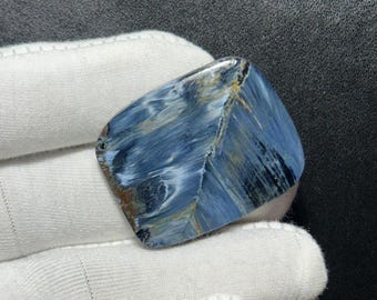 Könnte beinhalten: Ein polierter, unregelmäßig geformter Edelstein mit einem auffälligen blau-weißen Muster, akzentuiert durch goldene und braune Streifen. Die Oberfläche des Steins hat eine glatte, glänzende Oberfläche. Er wird von einer behandschuhten Hand gehalten.