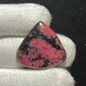 Puede incluir: Una gema triangular pulida con una vibrante paleta de colores. La piedra presenta una mezcla de rosa intenso, negro y toques de amarillo. La superficie pulida refleja la luz, resaltando los patrones y texturas únicos dentro de la piedra.