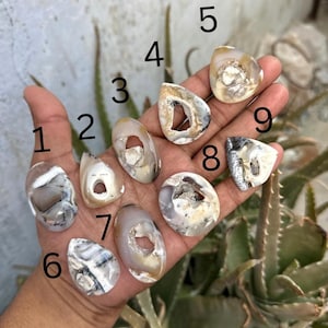 Può includere: Nove pietre d'agata levigate di forma irregolare con cavità centrali, numerate da uno a nove. Ogni pietra mostra bande uniche di colore bianco, grigio e marrone chiaro con texture cristalline all'interno dei centri cavi.