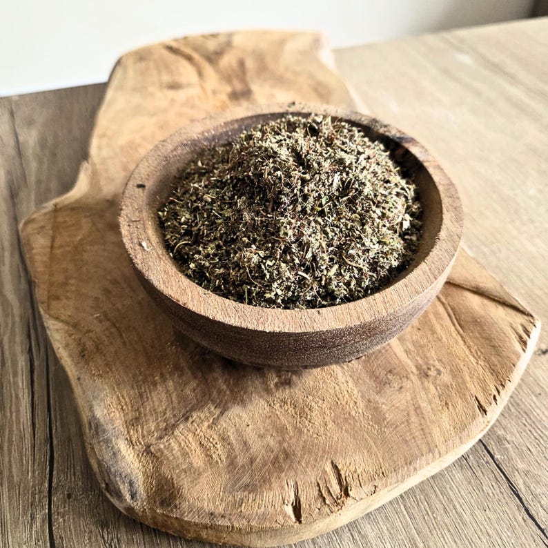 May include: A wooden bowl filled with dried herbs sits on a rustic wooden cutting board. The herbs are a mix of green and brown, and the bowl is a warm, natural wood tone. The cutting board has a unique, organic shape.