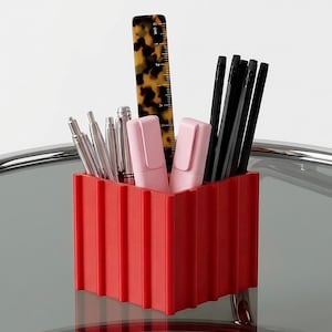 May include: A red desk tidy with a ridged design, containing various stationery items. The tidy holds silver pens, two pink highlighters, black pencils, and a ruler with a tortoiseshell pattern. The tidy sits on a glass surface.