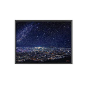 以下が含まれることがあります： 星空の下の夜の都市景観を描いたフレームプリント。画像は、多数の星と明るい天の川の帯で満たされた濃い青色の空を示しています。下の街の明かりが温かい輝きを生み出しています。