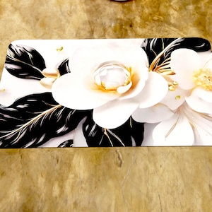 May include: Rectangular white bath mat with a large floral design. The flowers are white with gold accents, surrounded by black and gold leaves. The mat is designed for the floor.