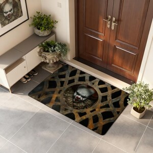May include: A rectangular doormat featuring a menacing clown face within a circular sewer grate design. The mat has a black background with a rusty, orange-brown metal grate pattern. The clown has white face paint, red lips, and a ruffled collar.