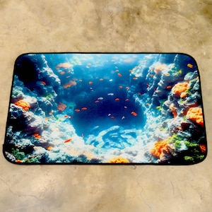 May include: Rectangular mat with an underwater scene. The design depicts a deep blue ocean with orange and white coral reefs. Many small orange and white fish are swimming. The mat has a black border. The mat's dimensions are in centimetres.