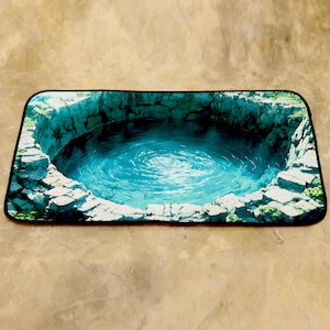 May include: Rectangular rug with a detailed illustration of a well. The well has clear blue water and stone walls. The rug has a black border. The well design creates a 3D effect. The rug is set against a neutral background.