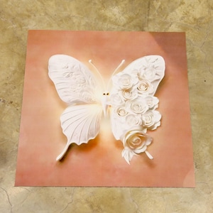 May include: A white butterfly wall art piece with intricate details. The butterfly's left wing features delicate patterns, while the right wing is adorned with white roses. The artwork is set against a soft, peach-coloured background.