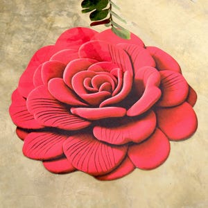 May include: A large, red, flower-shaped rug with detailed petal design. The rug is a vibrant shade of red, with darker shading to define the petals. A green leaf and stem are visible at the top. The rug is designed to resemble a rose.