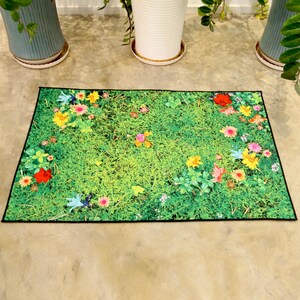 May include: A rectangular doormat with a vivid green grass pattern and colourful flowers. The mat has a black border and is on a concrete surface. The flowers include red, yellow, pink, and blue blooms.