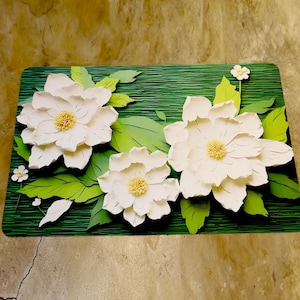 May include: A decorative floral piece with three large white flowers, yellow centres, and green leaves. The flowers are set on a textured green background, with small white flowers around the edges.