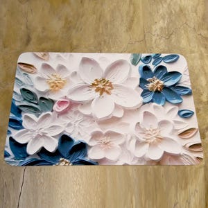 May include: A rectangular decorative mat with a 3D floral design. The flowers are in white, blue, and pink, with gold accents. The mat has a textured appearance, creating depth and dimension. Ideal for home decor.