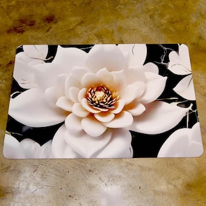 May include: Rectangular floral mat featuring a large, white flower with a golden centre against a black and white background. The flower has multiple layers of petals, creating a 3D effect. The mat is designed for indoor use.