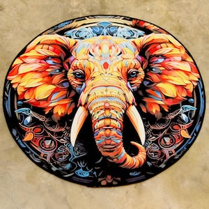 May include: A circular rug featuring a detailed elephant design. The elephant is depicted in vivid colours, including orange, yellow, and blue, set against a black backdrop with decorative patterns. The elephant's tusks are white.