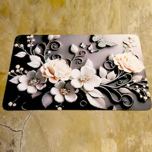 May include: Rectangular decorative mat with a floral design. It features white and peach-coloured flowers, grey leaves, and butterflies. The mat has a black and grey gradient background with a 3D effect.