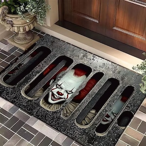 Op de afbeelding: Een rechthoekige deurmat met een eng clownontwerp. De mat heeft een donkergrijze, getextureerde achtergrond met zes rechthoekige uitsparingen. Elke uitsparing toont een deel van het gezicht van de clown, met rood haar, witte schmink en een sinistere grijns.